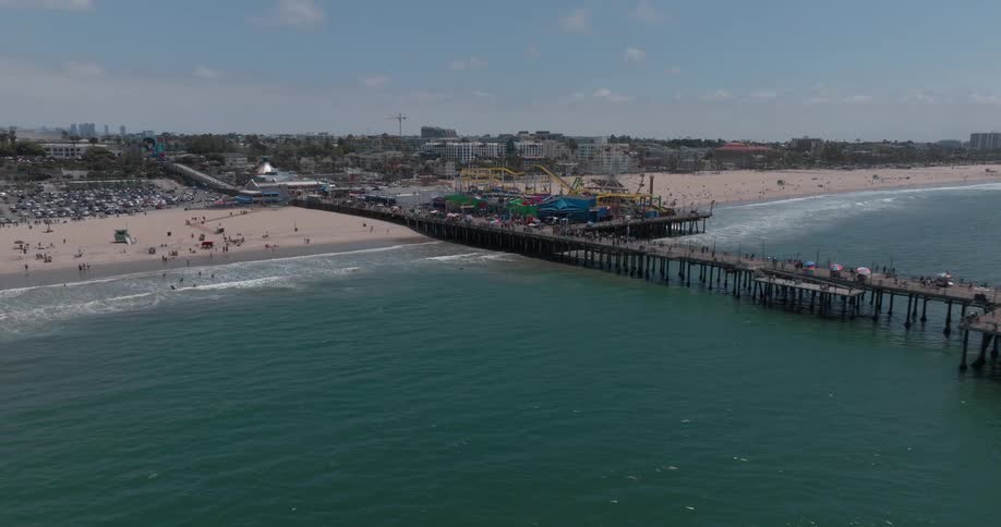 A large crowd of people gathers on the beach near an amusement park on the bay as ocean waves hit the shore with a city view in the background
