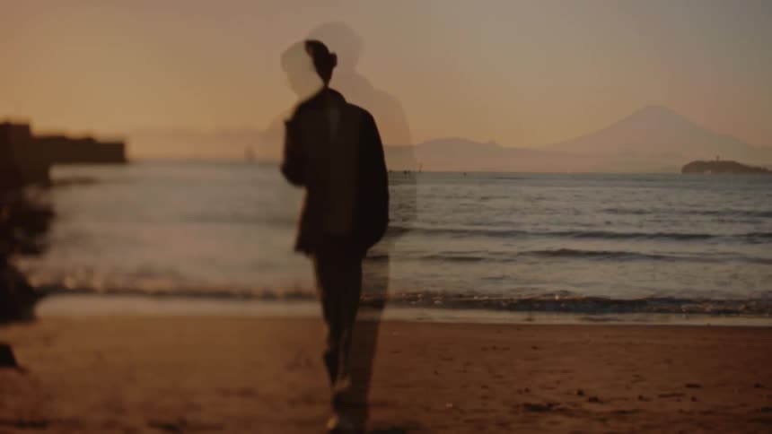 Person stands alone on a beach at sunset holding a phone as distant mountains form dark silhouettes along the glowing horizon