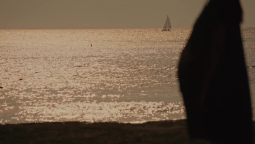 Silhouette of a person stands on the beach at sunset with a sailboat in the distance
