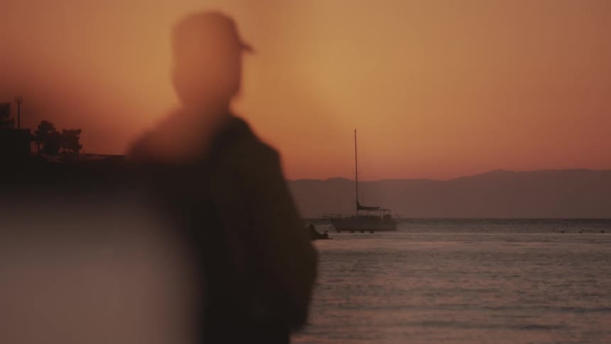 A silhouette person stands by the shore at sunset gazing toward a sailboat on the water