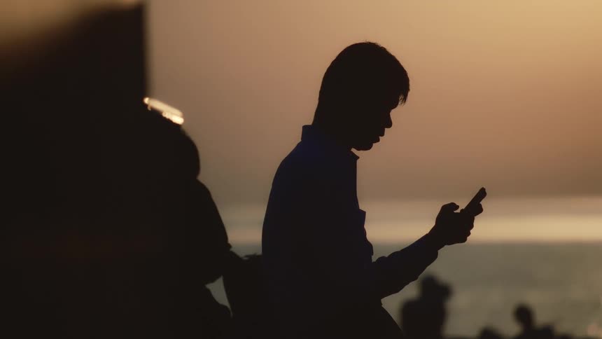 A man is silhouetted against a setting sun over water holding a cell phone in his hands
