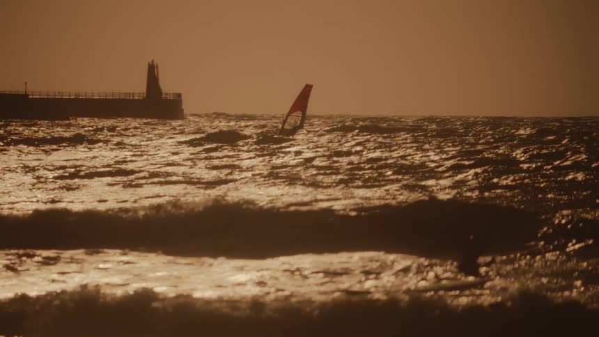 A windsurfer glides across shimmering waves near a pier at sunset