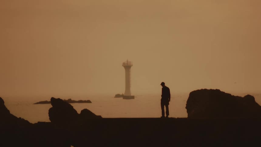 A silhouetted person stands on a rocky shore waving hands toward a lighthouse at sunset