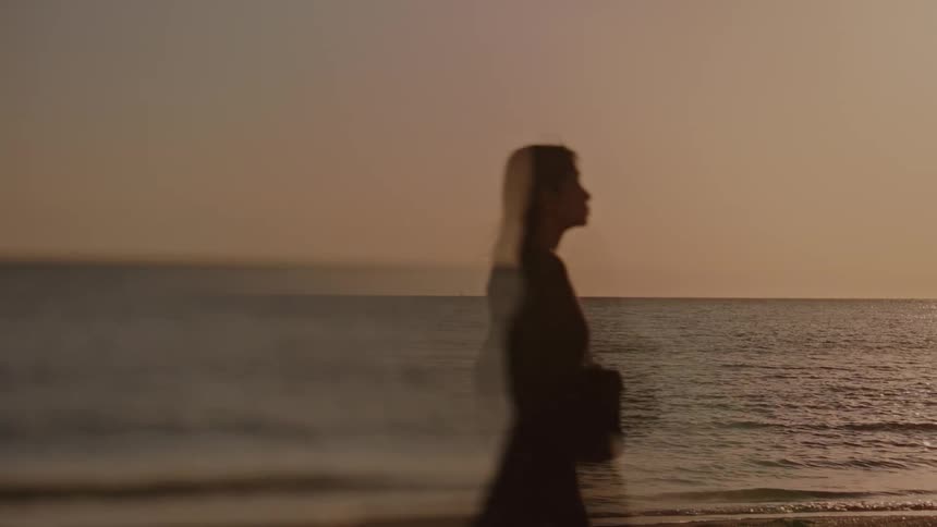 A woman walks along a beach at sunset carrying a shoulder bag with waves lapping at the shore