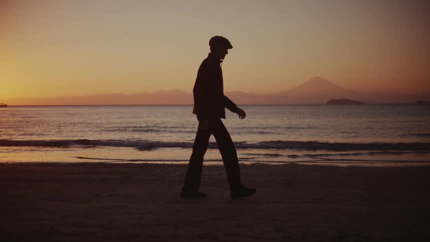 A person wearing a hat walks along the beach sand at sunset as ocean waves hit the shore