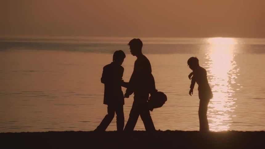 Silhouettes of kids throwing stones on a beach while another person walks during sunset
