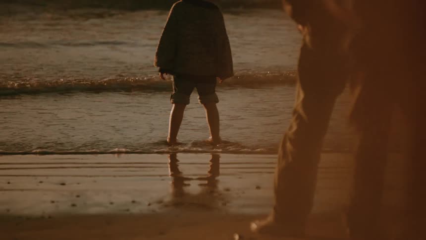 Child stands on a wet sandy beach at sunset as an adult watches from behind