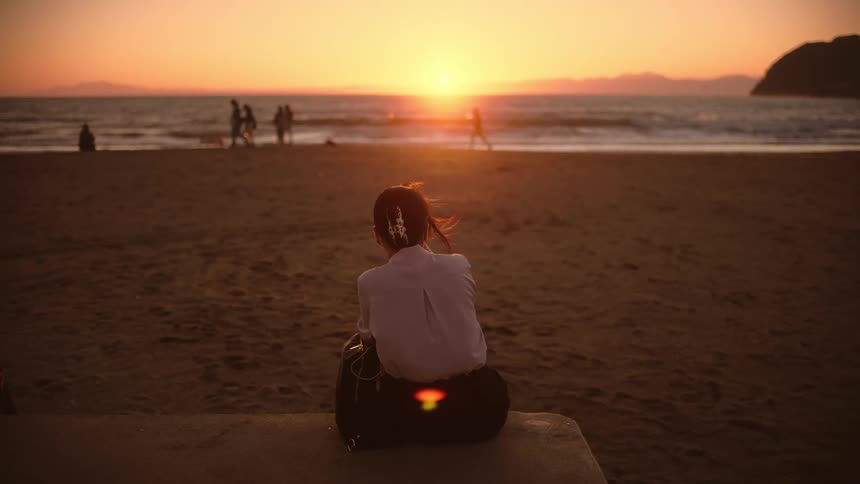 A woman sits on a bench at the beach watching children play along the shoreline as the sun sets