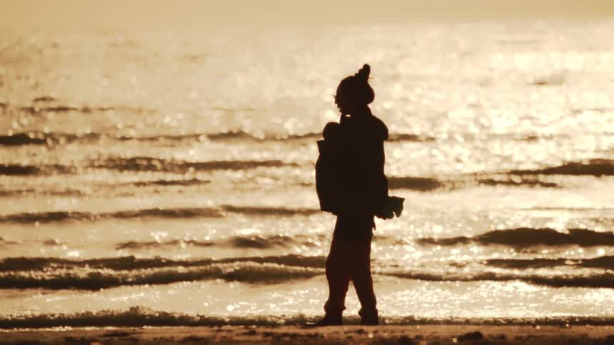 A person is silhouetted against a body of water with sunlight reflecting on the surface walking along the shoreline