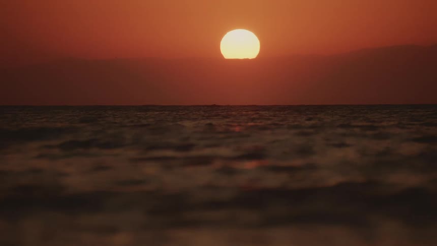 Sunset casts an orange glow across the ocean as the sun sinks behind distant clouds its light reflecting off the rippling water