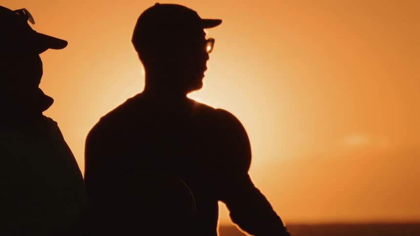 A man wearing glasses holds a table tennis paddle hitting the ball with the orange setting sun in the background