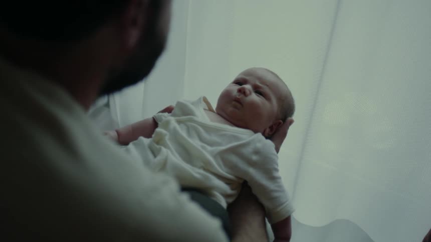 A man holds a baby near a sunlit window framed by white curtains
