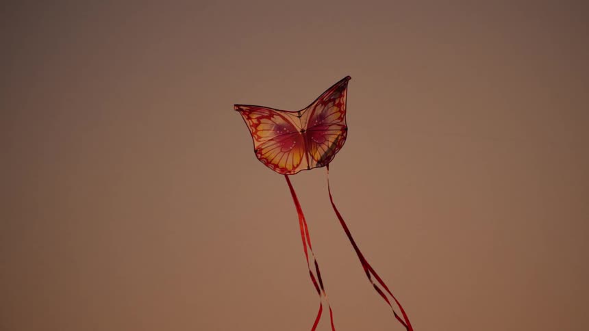 A butterflyshaped kite with red and white tails is flying against a gradient sky during sunset