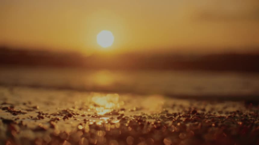 The sun sets over a body of water casting an orange glow and reflecting on the wet sand in the foreground