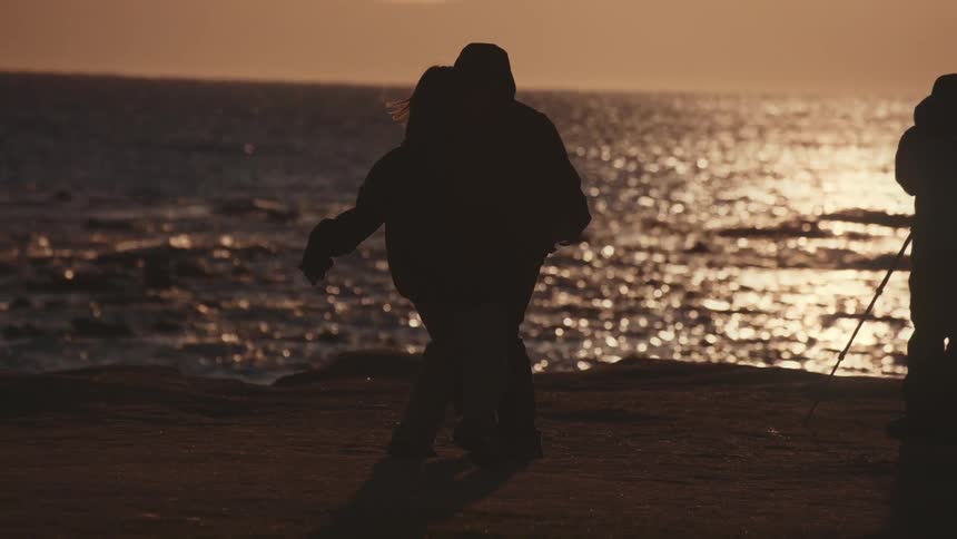 Two people play by the ocean at sunset as one person stands with a tripod