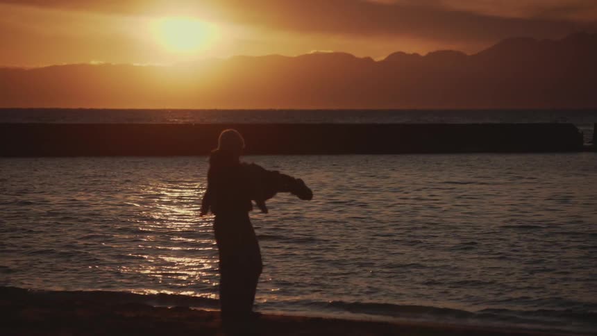 An adult carries a child on the beach spinning them in a 360degree turn creating fun and excitement as the orange setting sun appears in the background