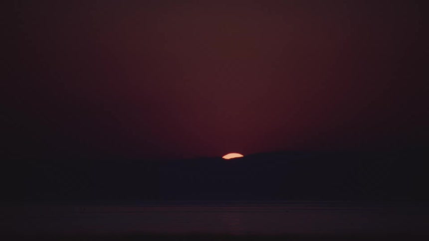 A timelapse shot captures the orange sun disappearing behind a silhouetted mountain range over a dark body of water