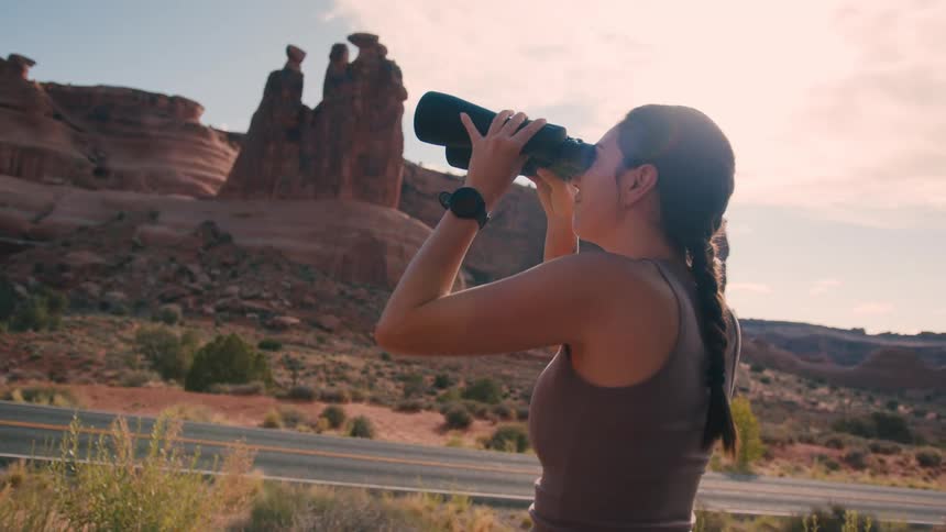 A woman uses binoculars to view distant red rock formations near a road in a sunlit desert landscape