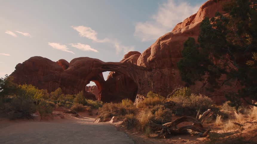 A natural sandstone arch rises from rocky terrain surrounded by desert shrubs under a partly cloudy sky
