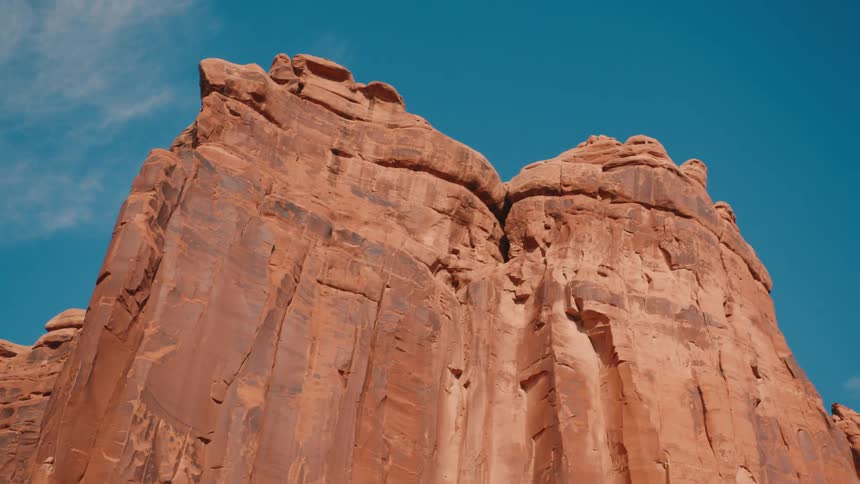 A towering red rock formation rises sharply against a clear blue sky surrounded by rugged desert terrain