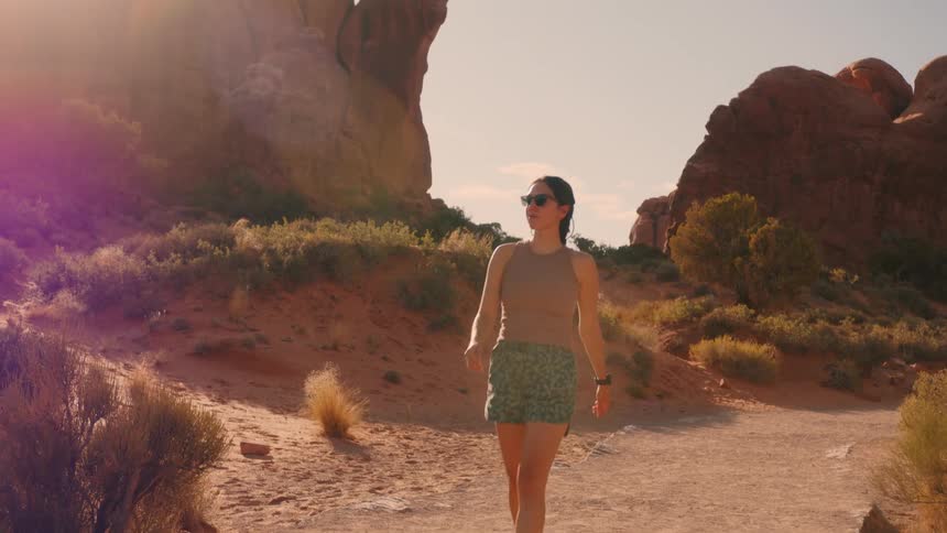 A woman wearing sunglasses is walking on a path in a desert and she is wearing shorts and a black boot