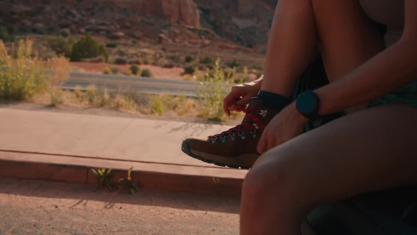 A young woman sits in the trunk of an suv tying her shoelaces while parked on the side of the road surrounded by mountains at midday