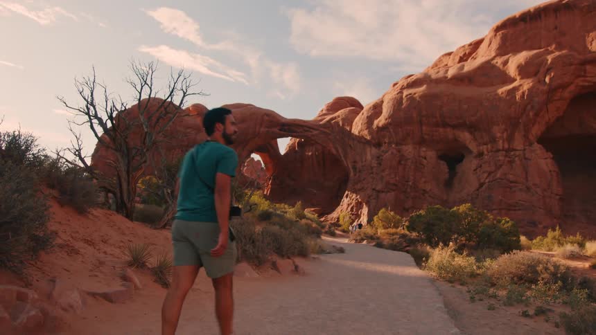 A man hikes through a desert landscape toward a natural red rock arch formation