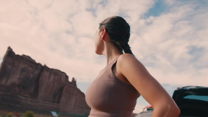 A young woman stands up from the trunk of her car after tying her shoelaces smiling as she takes in the view of the surrounding rocks and mountains