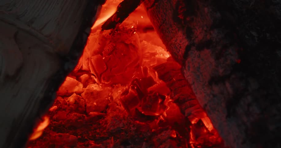 Glowing embers and charred wood crackle inside a firepit radiating intense orange and red heat