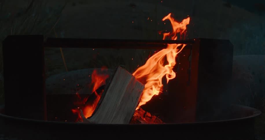 Flames engulf a piece of wood inside a round metal fire pit casting a warm orange glow amid dark surroundings