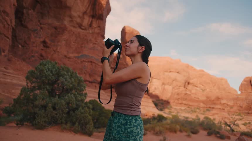 A woman standing in a desert taking pictures of a mountain of reddish rocks