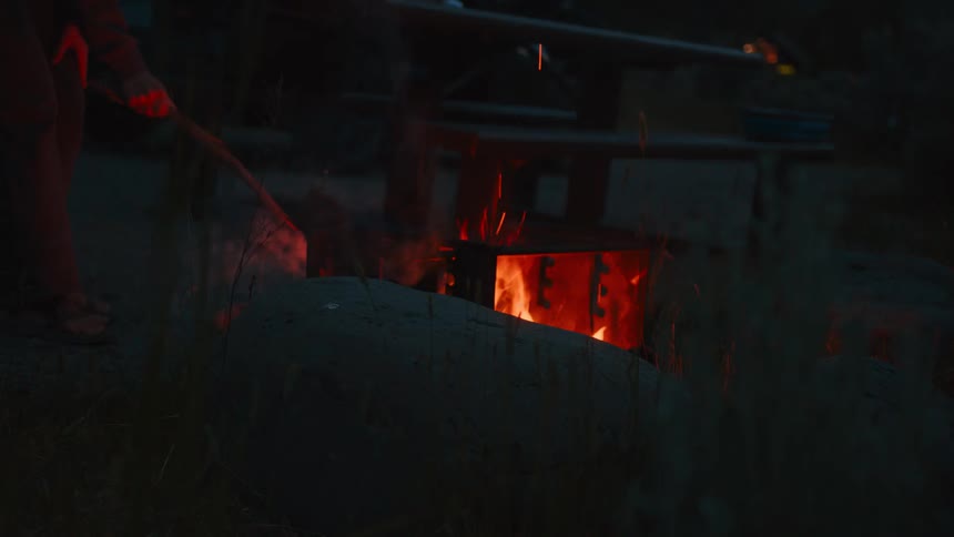 A person uses a stick to spread burning wood in an outdoor fireplace at night