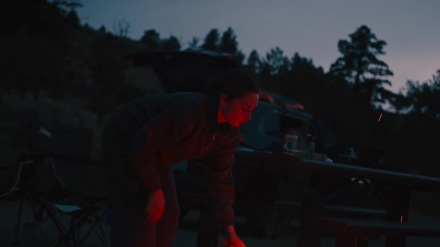 A young woman sets up a campfire in the woods as evening turns to night