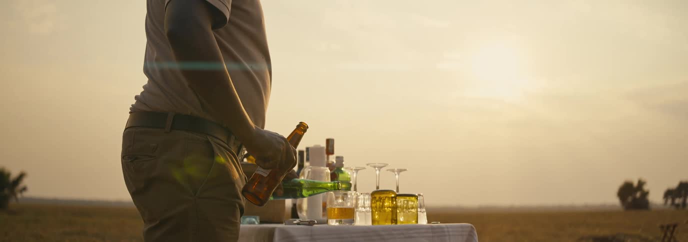 A man pours a drink into a glass on an outdoor table in a grassland on a sunny midday