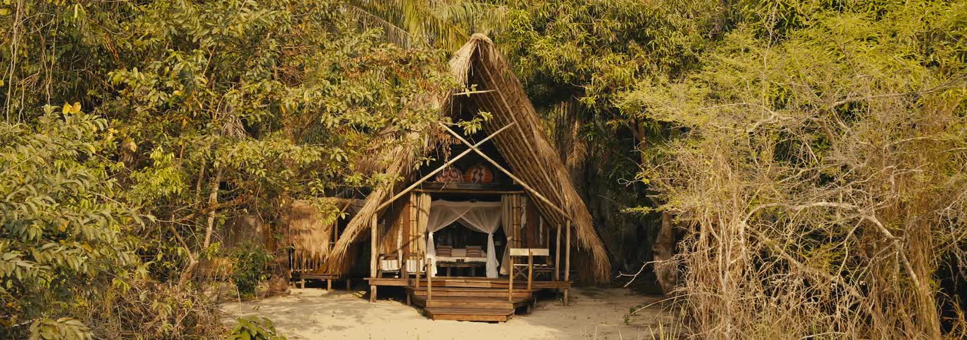 A thatched roof hut sits nestled in dense tropical forest just off a sandy clearing