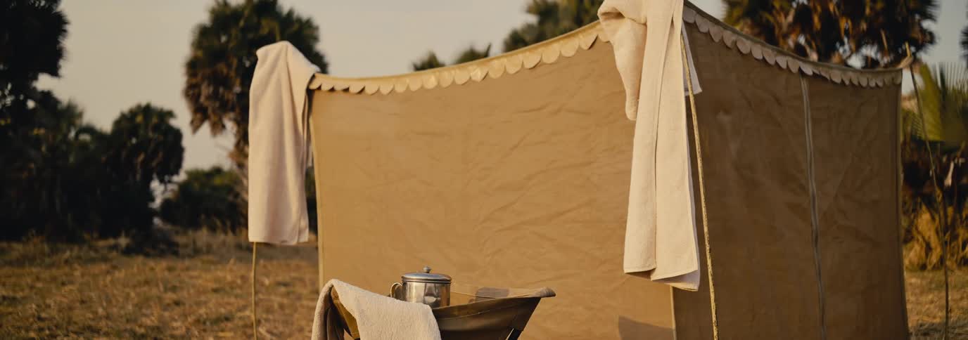 A beige canvas tent stands on dry grass with towels draped on its sides and a metal pot resting on a wooden table out front