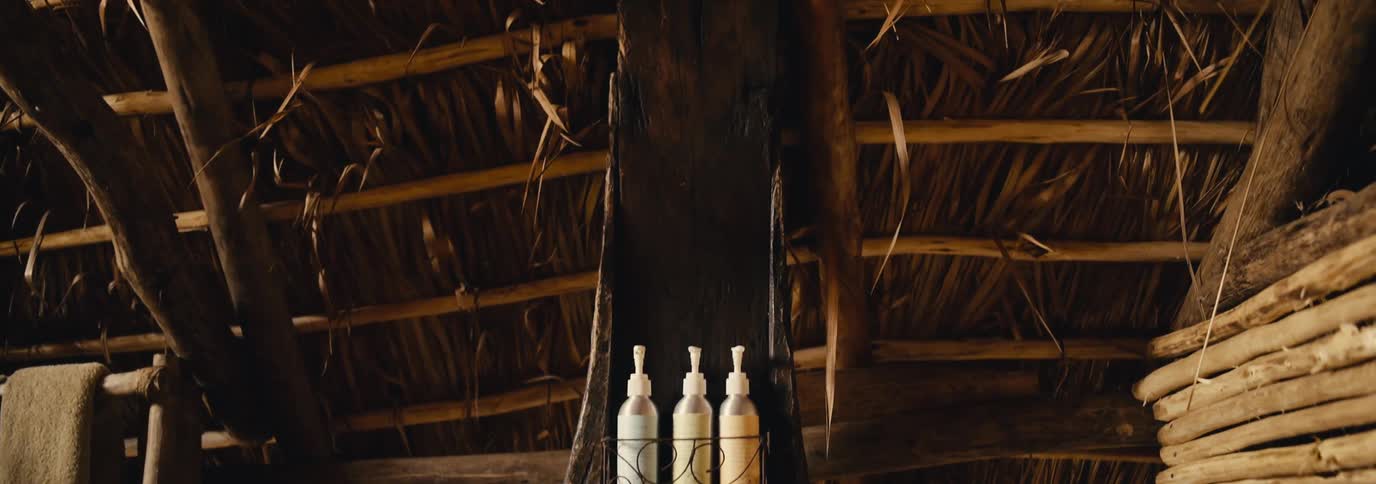 A showerhead hangs from wooden support beams and dried palm leaves in a rustic thatchedroof bathroom