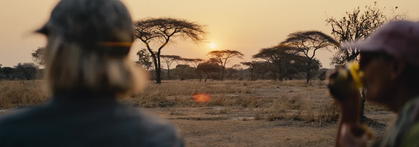 A man and a woman sit side by side as the man drinks from a glass while they both gaze through the safari jungle at sunset