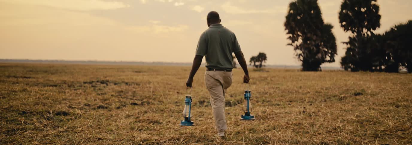 A black man walks through a grassland in the wild at midday holding two lamps