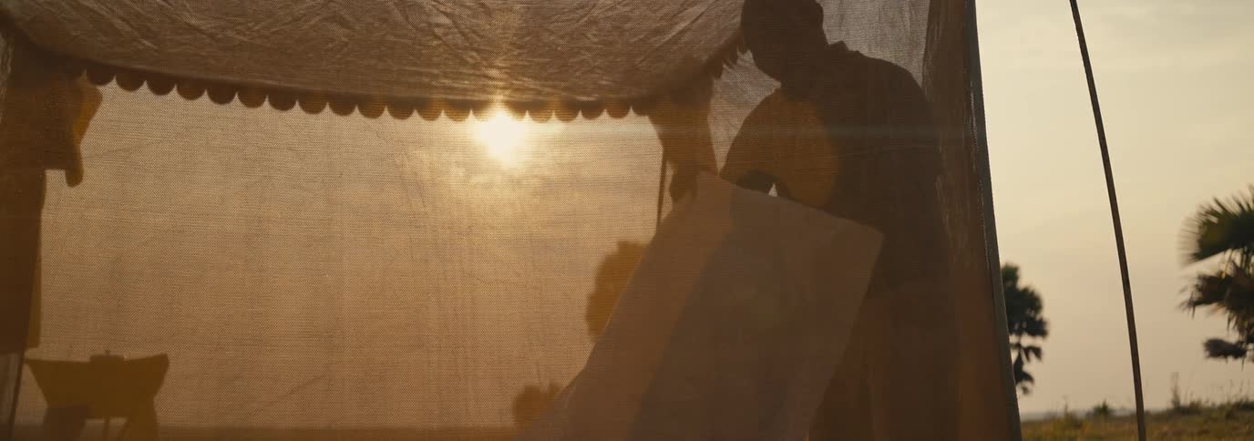 A person sets up a tent in a grassy field at sunset with the silhouettes of a chair and trees visible through the tents fabric