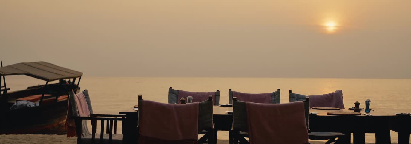 A serene sunset over the ocean with a dining setup featuring wooden chairs draped with pink blankets