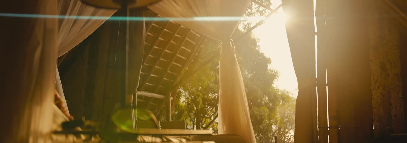 Soft sunlight filters through flowing white curtains in a rustic openair room with wooden beams and a view of trees outside