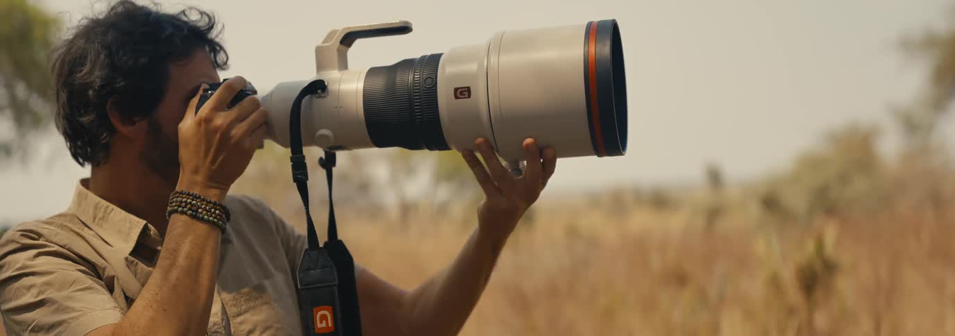 A person in a lightcolored shirt and beaded bracelet looks through the viewfinder of a camera with an oversized white and black telephoto lens