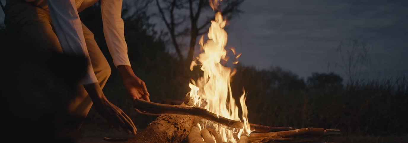 A black man adding wood to a fire outdoors