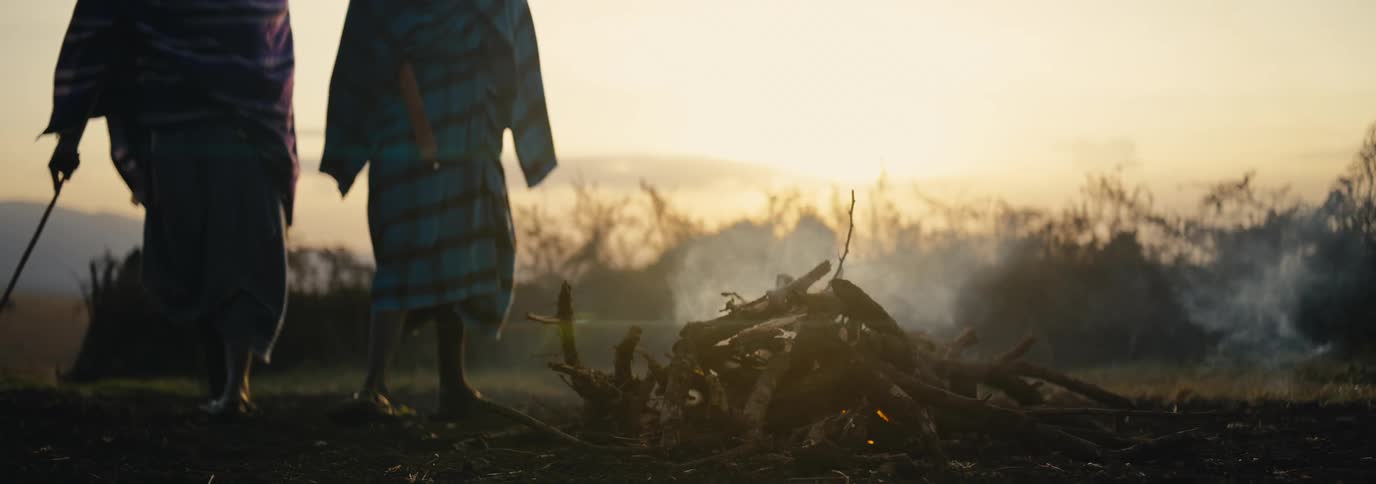 Two people stand near a smoking fire in a field under a red tinted sky