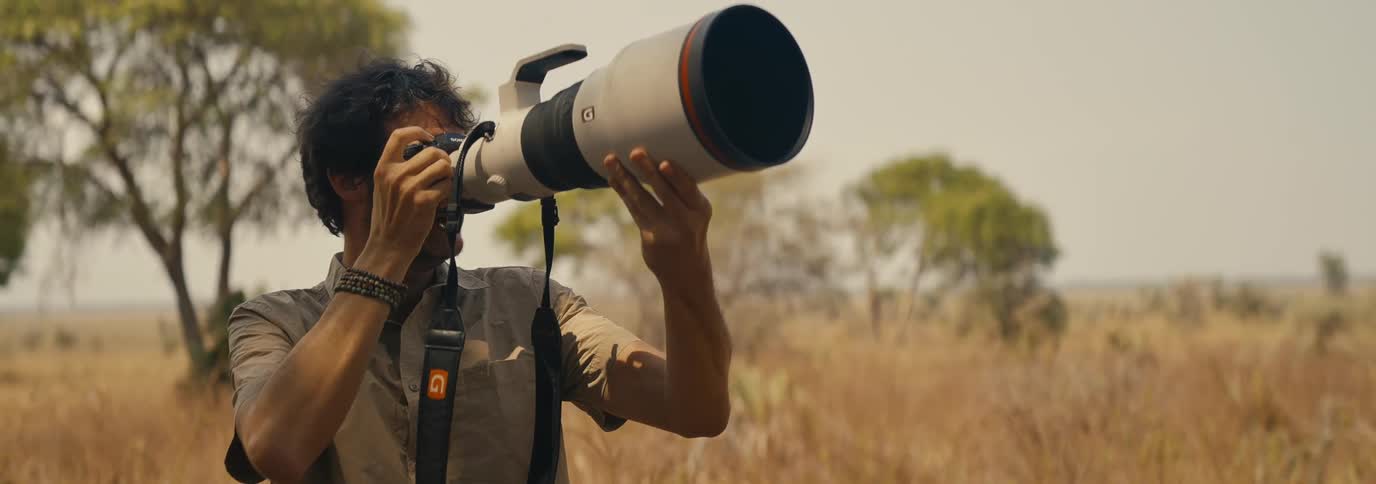 A wildlife photographer aims a large telephoto lens camera while standing in a dry grassy plain