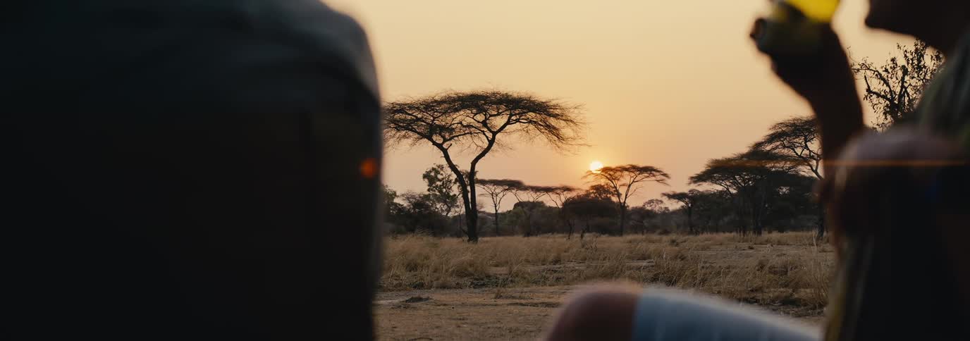Two people sit outdoors at sunset one holding a drink with silhouetted acacia trees in the background