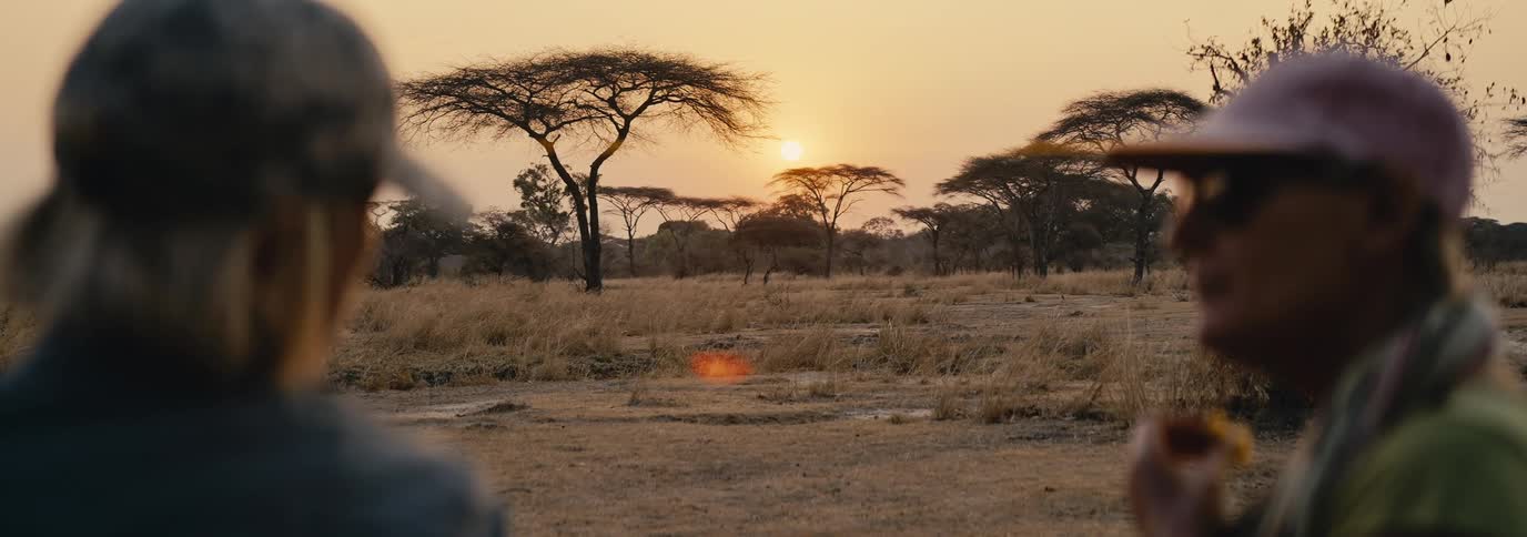 A young woman and a man both wearing baseball hats are in the wild with the setting sun visible in the distance