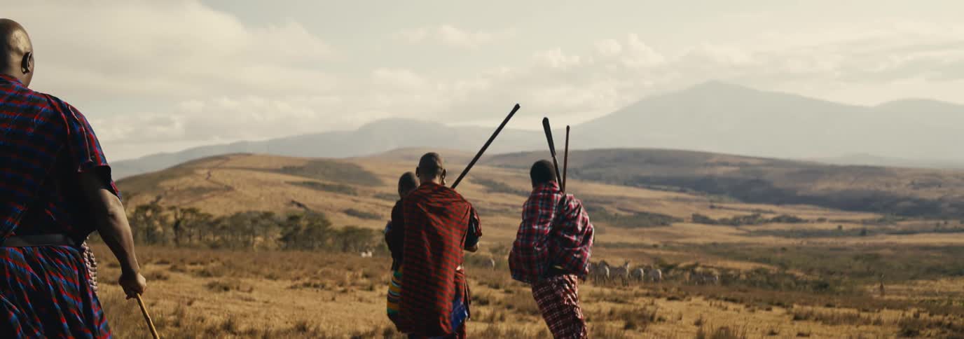Four tribal maasai people dressed in patterned fabrics and carrying sticks walk through a vast dry landscape with rolling hills and scattered trees under a light sky