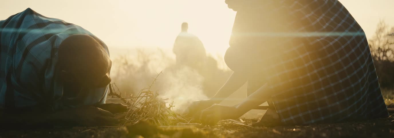 Two people kneel over a straw bundle in a sunlit outdoor space one blowing gently to kindle a small flame